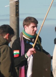 Picture of Frankie Rufolo at the protest. He's wearing a black jacket covered in patches, a coloured scarf, and a flag-pole in his right hand