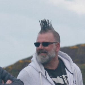 Man with spiky hair and dark sunglasses and a dark grey beard