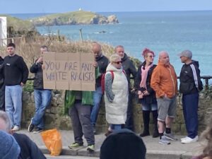 Large cardboard sign "you are anti white racists". Some people milling about behind, and a see view in the background
