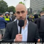 Nick Tenconi speaking into a microphone with Plymouth Civic Centre in the background. He is wearing a white shirt and black jacket. Skinhead with a short beard and mustache